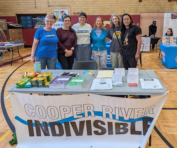 We are in a school gym. In the foreground is a white folding table with the Cooper River Indivisible banner. On the table is school supplies, flyers, and cards. Behind the table are six people arranged from left to right and they are shoulder to shoulder