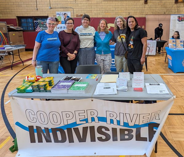 We are in a school gym. In the foreground is a white folding table with the Cooper River Indivisible banner. On the table is school supplies, flyers, and cards. Behind the table are six people arranged from left to right and they are shoulder to shoulder