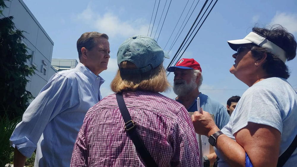 Four people stand around in circle talking about the state of the world. On the far left of the picture (but not far enough left on the political spectrum) is Donald Norcross. He is wearing a blue button down shirt with no tie or jacket. In the middle of the picture we see the back of a person and across from them, a person in a Phillies hat. Finally, on the right is a women wearing a white visor