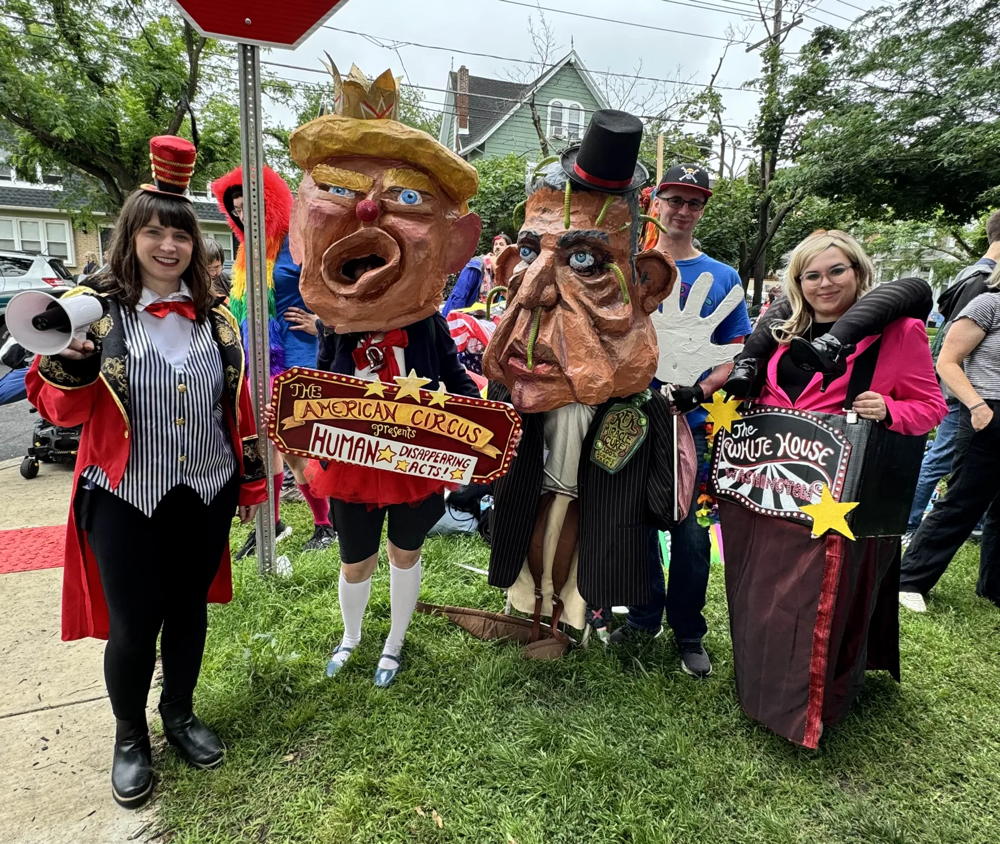 The Creative Team stands side by side under a stop sign. The person on the left is dressed like a ring master for a circus. The person to the right of the ring master is a caricature of Donald Trump and they have a sign that says, "The American Circus Presents Human Disappearance Acts!" The a person is a caricature of RFK Jr. and finally a person with a pink blazer has a sign that says "The White House"