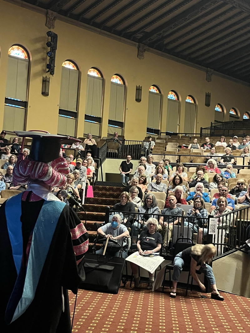 It is a picture of our keynote speaker for the June meeting taken from behind. The speaker is in drag and is dressed like a college graduate. They are standing in front of a microphone and we can see the left side of the auditorium. There are a series of high windows and row after row of people in attendance
