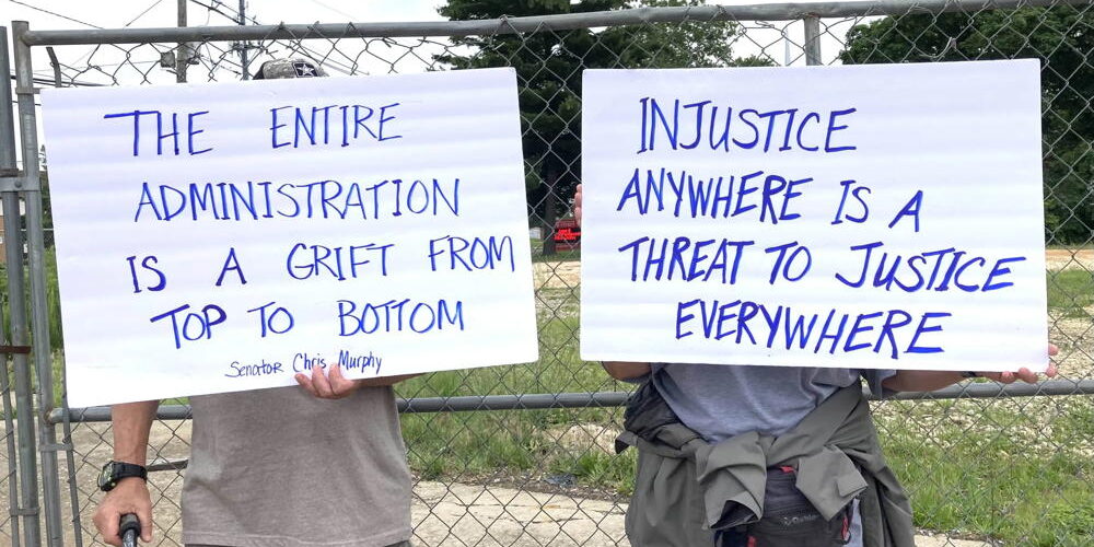 Two protestors stand in front of a metal fence and hold up signs. The one the left says, "The Entire Administration is a Grift from Top to Bottom" and the one the right says, "Injustice Anywhere is the Threat to Justice Everywhere"