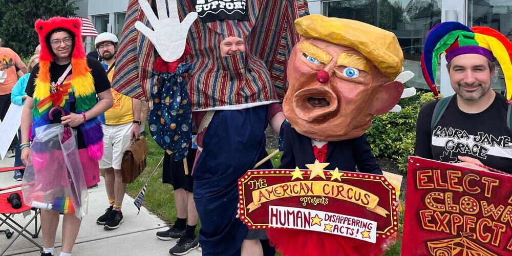 Four members of the CRI Creative Team stand are protesting in front of a Tesla Dealership. On the right the person is wearing a jester hat and holding a sign that says, "Elect a Clown, Expect a Circus". The next person is wearing a Donald Trump disguise and they are holding a sign that says, "The American Circus Presents Human Disappearing Acts". Then we see the bottom half of a puppet mocking Elon Musk and finally a person wearing a rainbow hat that comes down to their waist