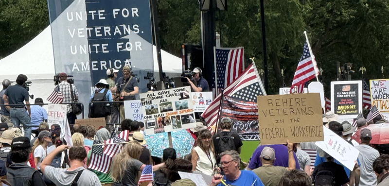 A protest in DC. A group of people is milling about under a banner that says "Unite for Veterans. Unite for America". Some of the people are holding signs including one that says "Rehire Veterans in the Federal Workforce" and others have American Flags