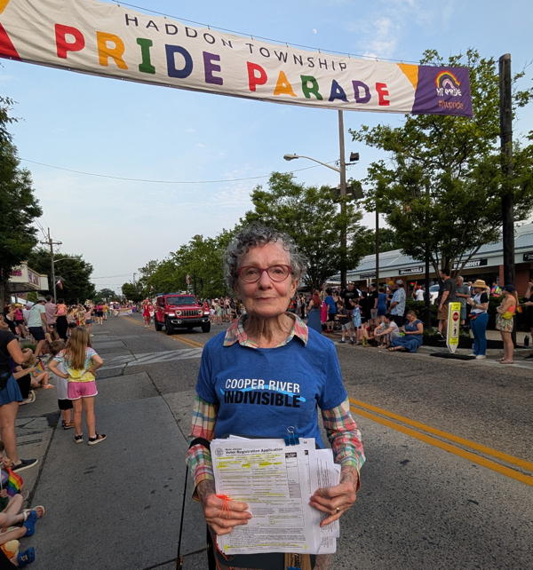 A woman in a blue cooper river indivisible shirt stands in the street under a banner that says "Haddon Township Pride Parade". She is holding a clipboard with voter registration forms