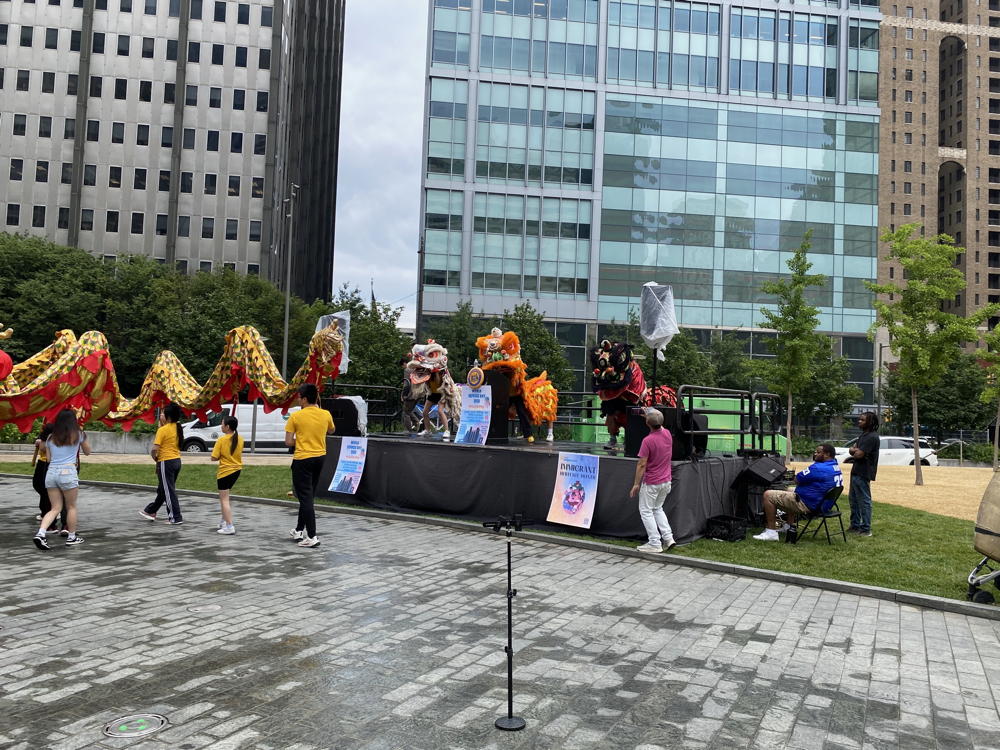 A stage is set up in Love Park in Philadelphia. On the stage are three small dragon creatures being manned by teenagers. In front of the stage are a whole bunch of people working a much larger dragon