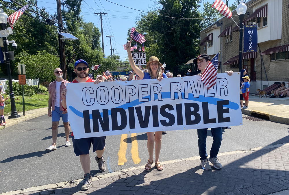 Three people are marching towards the camera holding the Cooper River Indivisible Banner. The person in the middle is enthusiastically waving the American Flag.
