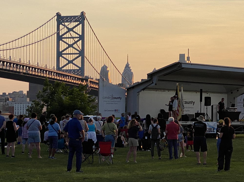 A crowd of people stand in front of a stage as the sun sets in the background. The sky is orange and we can see some sun glittering of sky scrapers in the background. On the left side of the image the bridge stretches across the river