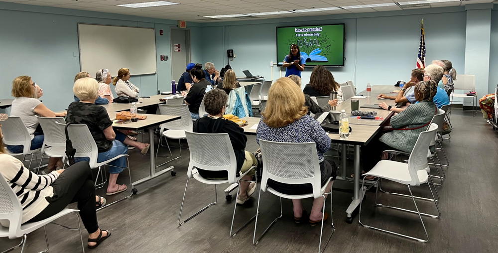 A series of tables form a rectangle and people are sitting all around it watching the teacher present their lesson. A screen in the front is used to display the computer slideshow for the people learning