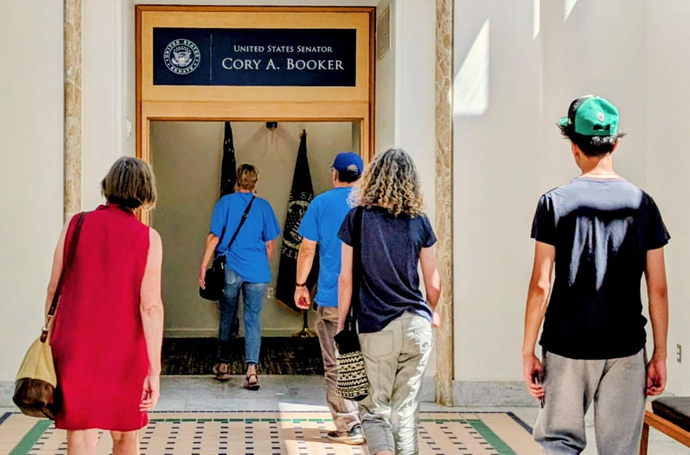 A group of badass CRI members strut into the offices of Cory Booker to talk about ways to support families of immigrant detainees. On the far left (farther left than Bernie Sanders) we have a woman in a red dress . In the middle and farthest from the camera are two people in the blue shirt of Cooper River Indivisible. On the right (physically, not politically) are a mother and her adult son.